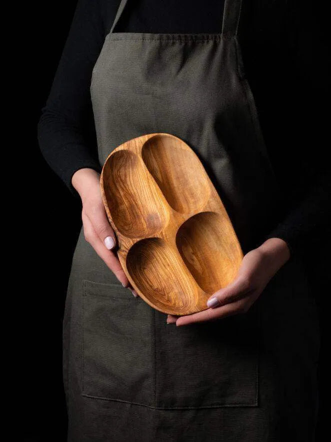 Handmade darido olive wood snack plate with four smooth compartments held by person in dark apron. - Handmade Olive Wood Snack Plate – 4 Compartments | Elegant, Eco-Friendly & Unique