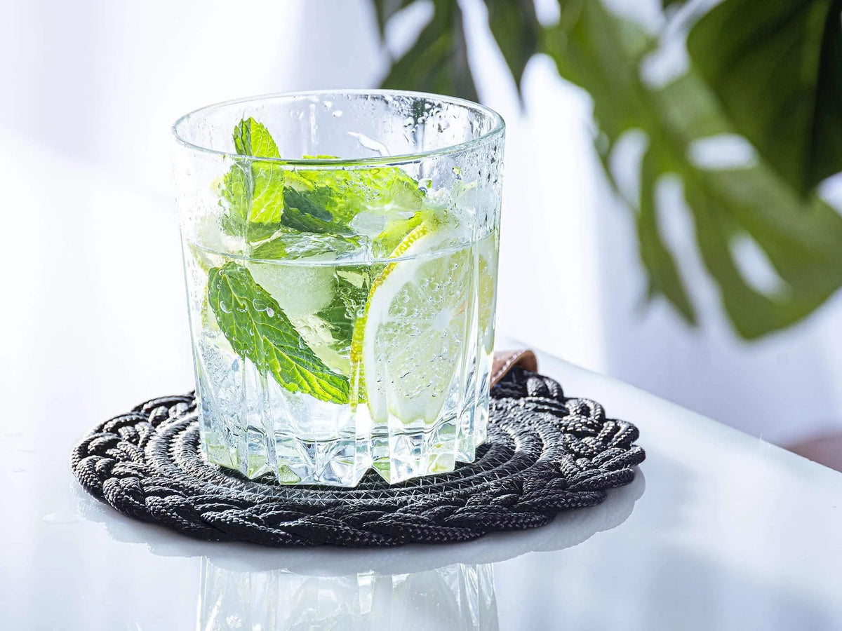 Set of 2 black cotton Darido coasters with intricate braided design under a glass of lime and mint drink. - Set of 2 Black Coasters- 15cm